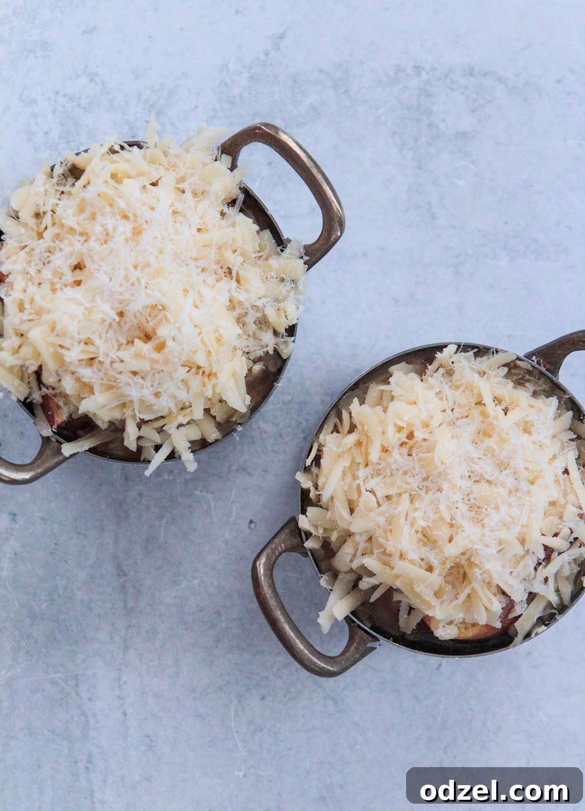 Savory Caramelized Onion Gratin 6 Two ceramic French onion soup bowls topped with a generous layer of shredded white cheese, ready for the broiler.