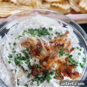 Potato chips surrounding a clear bowl of French onion dip, garnished with chives and caramelized onions.