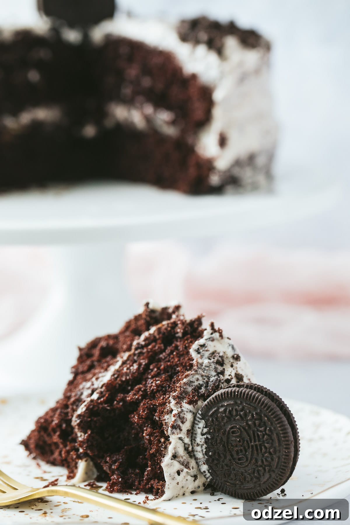 A perfectly sliced piece of Oreo cake on a pristine white plate, accompanied by a gold fork. In the softly blurred background, the remaining Oreo cake rests elegantly on a cake stand.