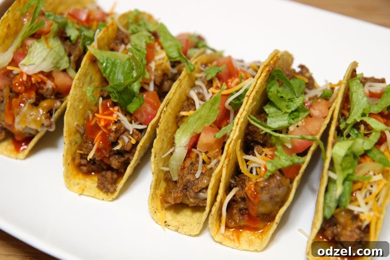 Close-up of crispy shell beef tacos showcasing melted cheese, diced tomatoes, and shredded lettuce on a white plate.