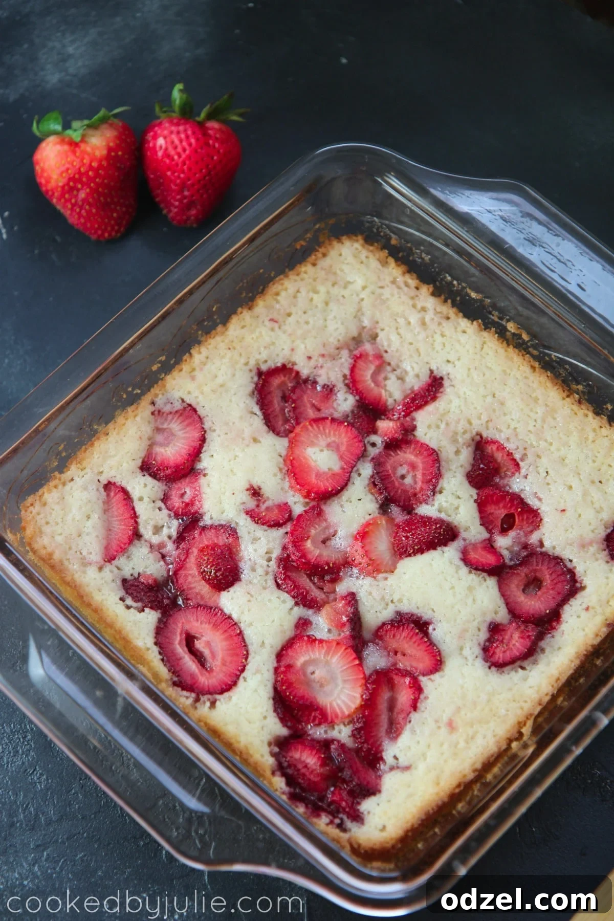 A close-up shot of baked strawberry cobbler in a glass baking dish, sitting on a dark surface with two fresh strawberries nearby, highlighting its golden crust and juicy filling.