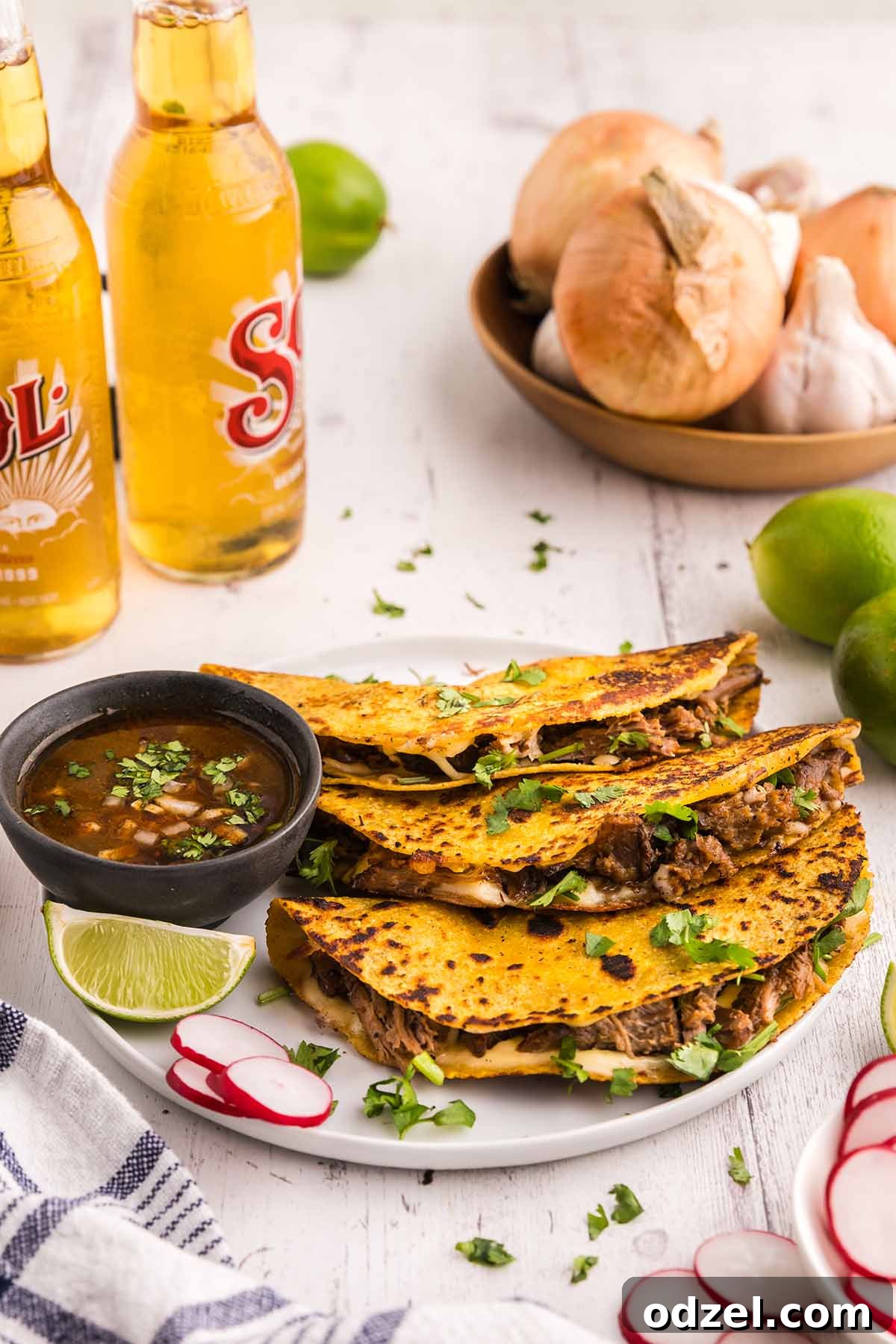 Three vibrant Birria tacos, garnished with fresh cilantro and diced white onion, beautifully arranged next to small bowls of rich consomé, lime wedges, and refreshing sodas.