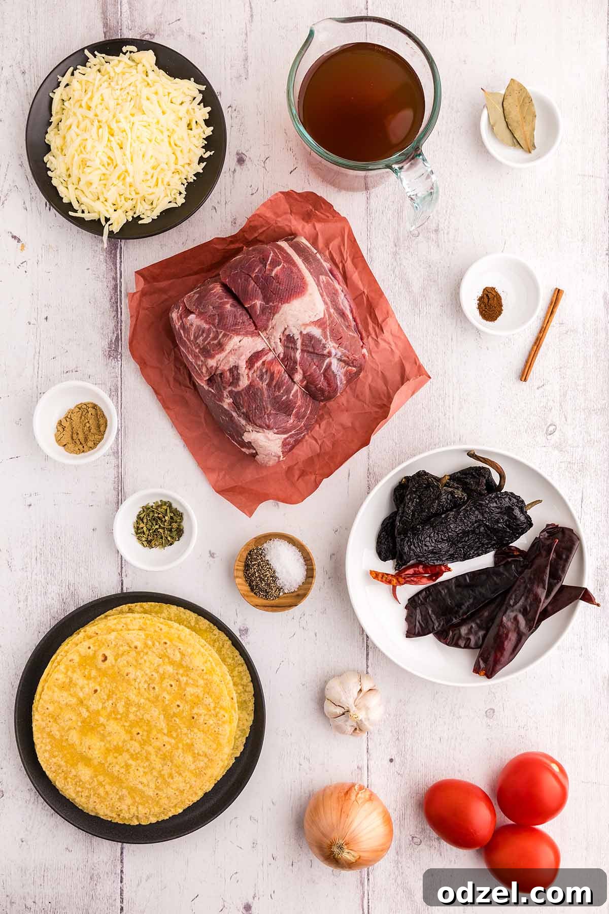 A vibrant flat lay of fresh ingredients for Birria Tacos: a raw beef chuck roast, a variety of dried chilies, fresh roma tomatoes, a white onion, garlic cloves, an assortment of spices including cumin, oregano, and ground cloves, a cinnamon stick, bay leaves, beef broth, a stack of corn tortillas, and shredded Oaxaca cheese on a white surface.