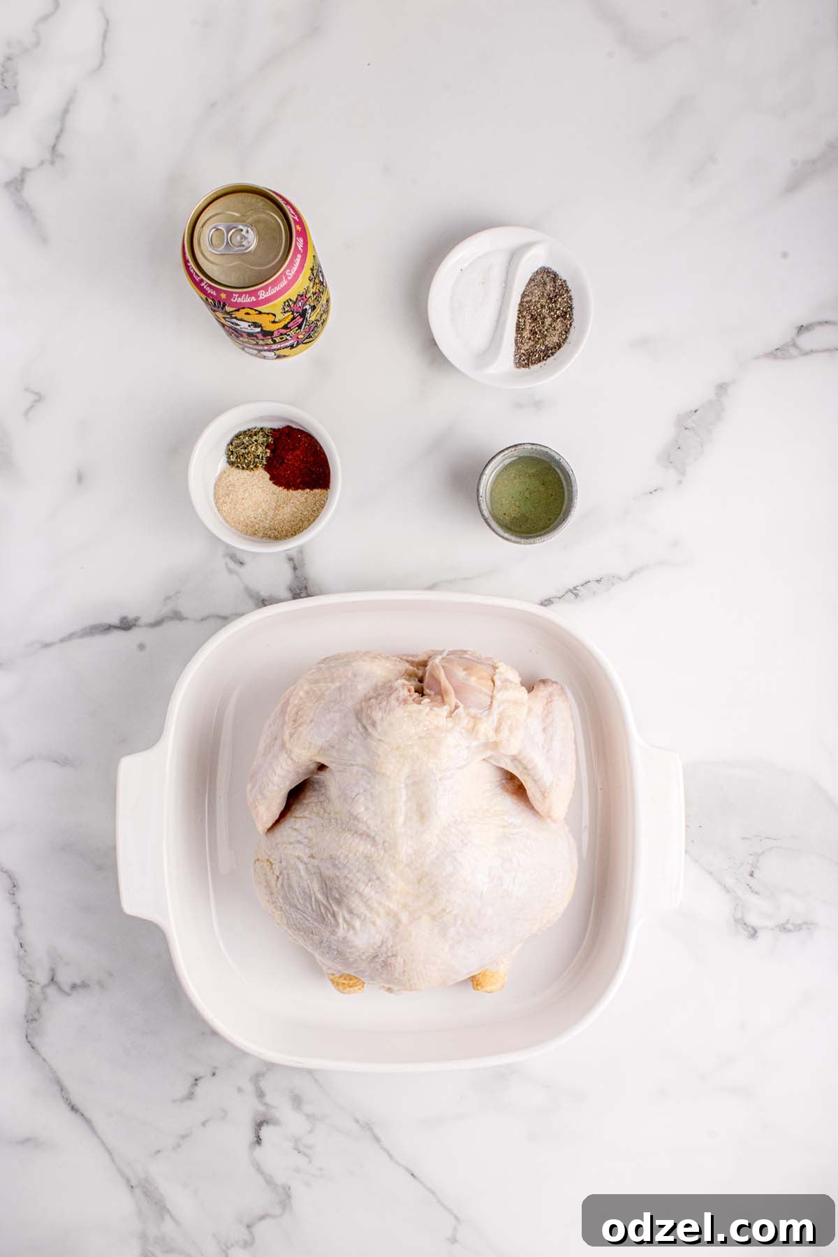 A raw chicken, a selection of fresh spices, a beer can, and a bottle of olive oil arranged on a rustic surface.