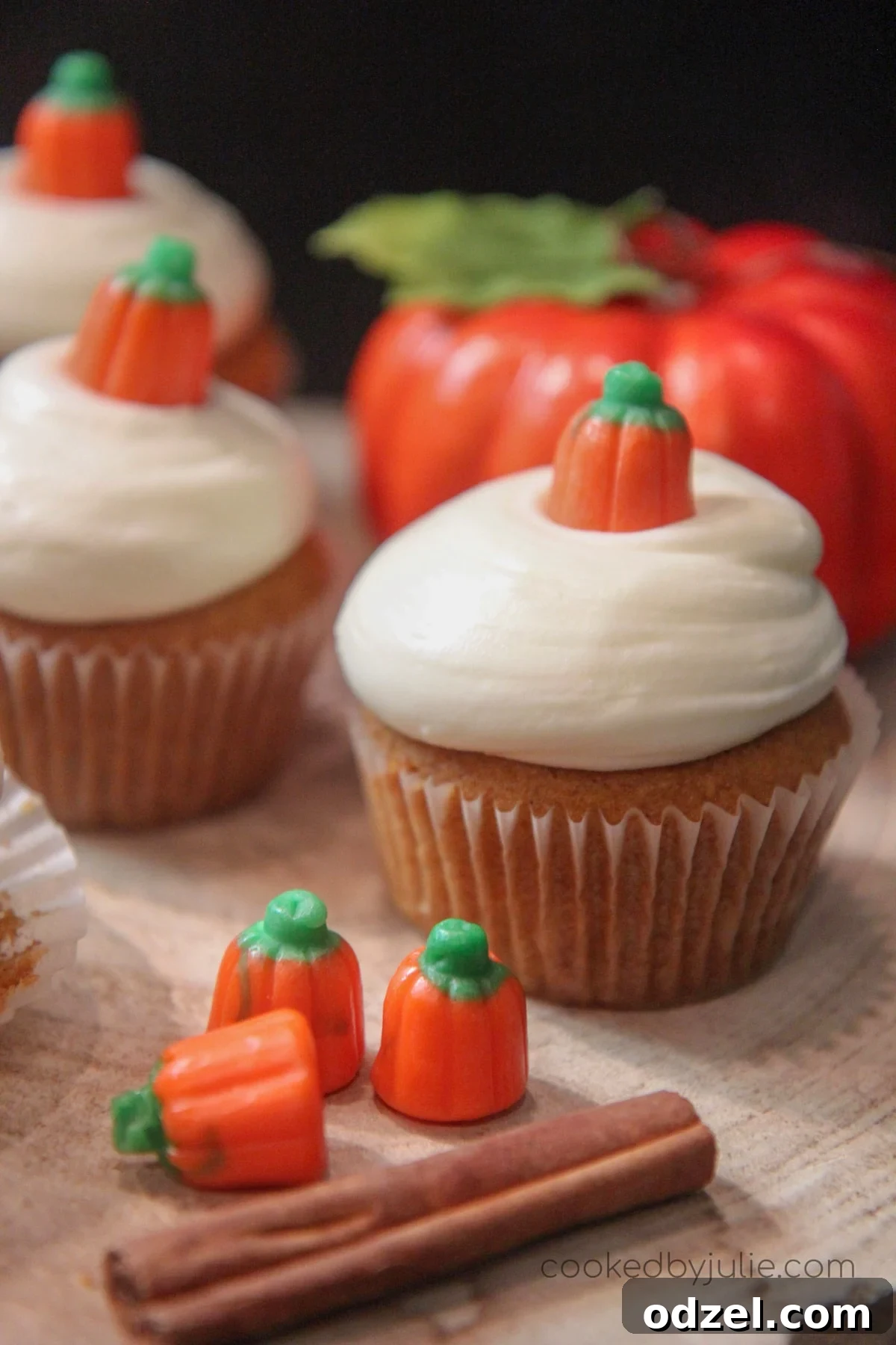 Three perfectly frosted pumpkin cupcakes, garnished with a cinnamon stick and cute pumpkin candies, sitting beside a decorative fake pumpkin, evoking a cozy fall atmosphere.