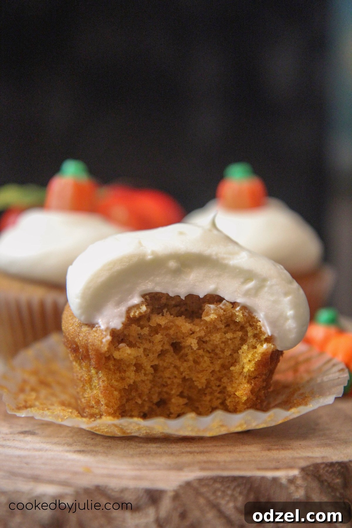 A deliciously moist pumpkin cupcake sliced in half, revealing its tender, spiced interior, artfully arranged on a wooden board with two whole frosted cupcakes in the soft-focus background, highlighting the perfect crumb.