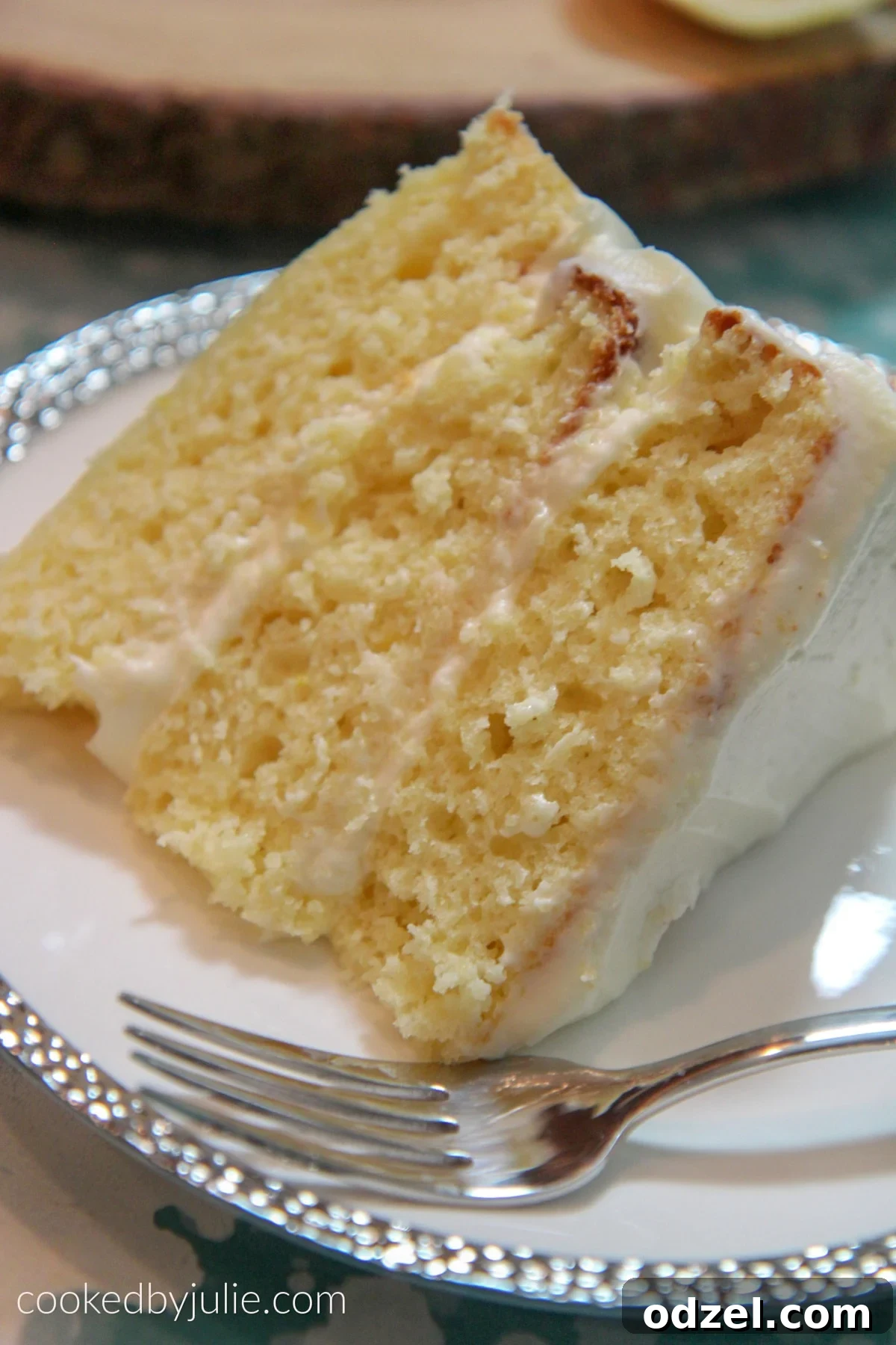 A perfectly sliced three-layer lemon cake on a white and silver plate with a fork beside it, showcasing the moist crumb and generous frosting.