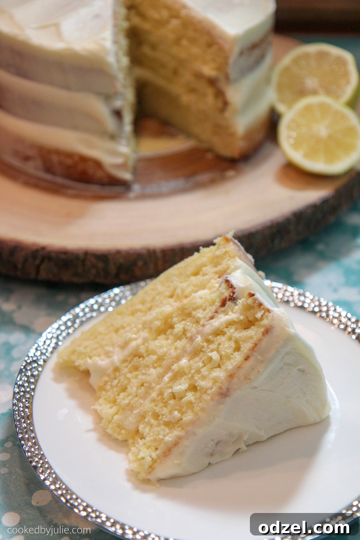 A delightful lemon cake slice on a white and silver plate, with the full lemon cake adorned with two fresh lemon slices in the background on a wooden stand.