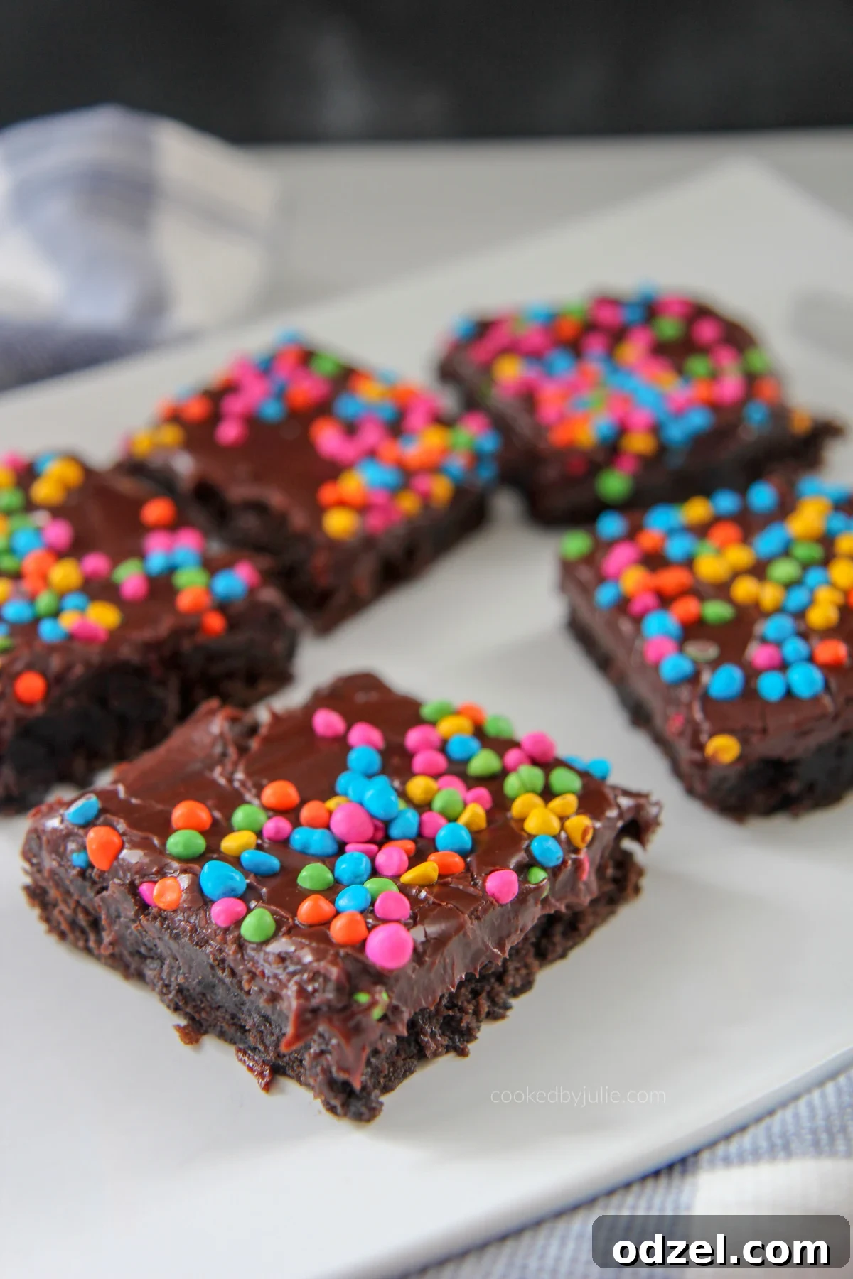 Dark Matter Brownies 2 A perfectly baked cosmic brownie topped with colorful rainbow chips, glistening with chocolate ganache, served on a pristine white plate.