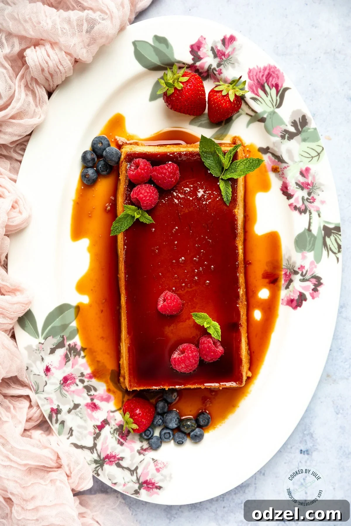 A perfectly prepared flan with a glossy caramel topping, garnished with fresh raspberries, blueberries, and mint leaves, presented on a white serving platter next to a soft pink linen.