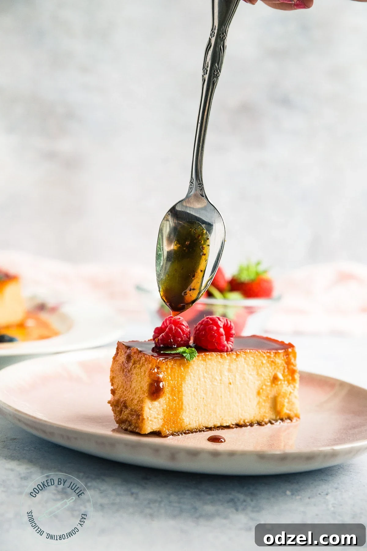 A delicious slice of caramel flan on a pink plate, with a spoon gently drizzling extra caramel sauce over the top, highlighting its rich, glossy texture.