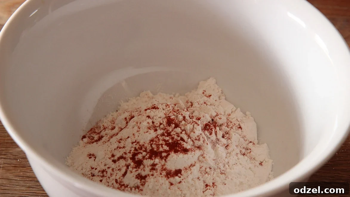 A white bowl holds all-purpose flour, surrounded by small piles of paprika, granulated garlic, oregano, salt, and pepper, representing the seasoned flour mixture.
