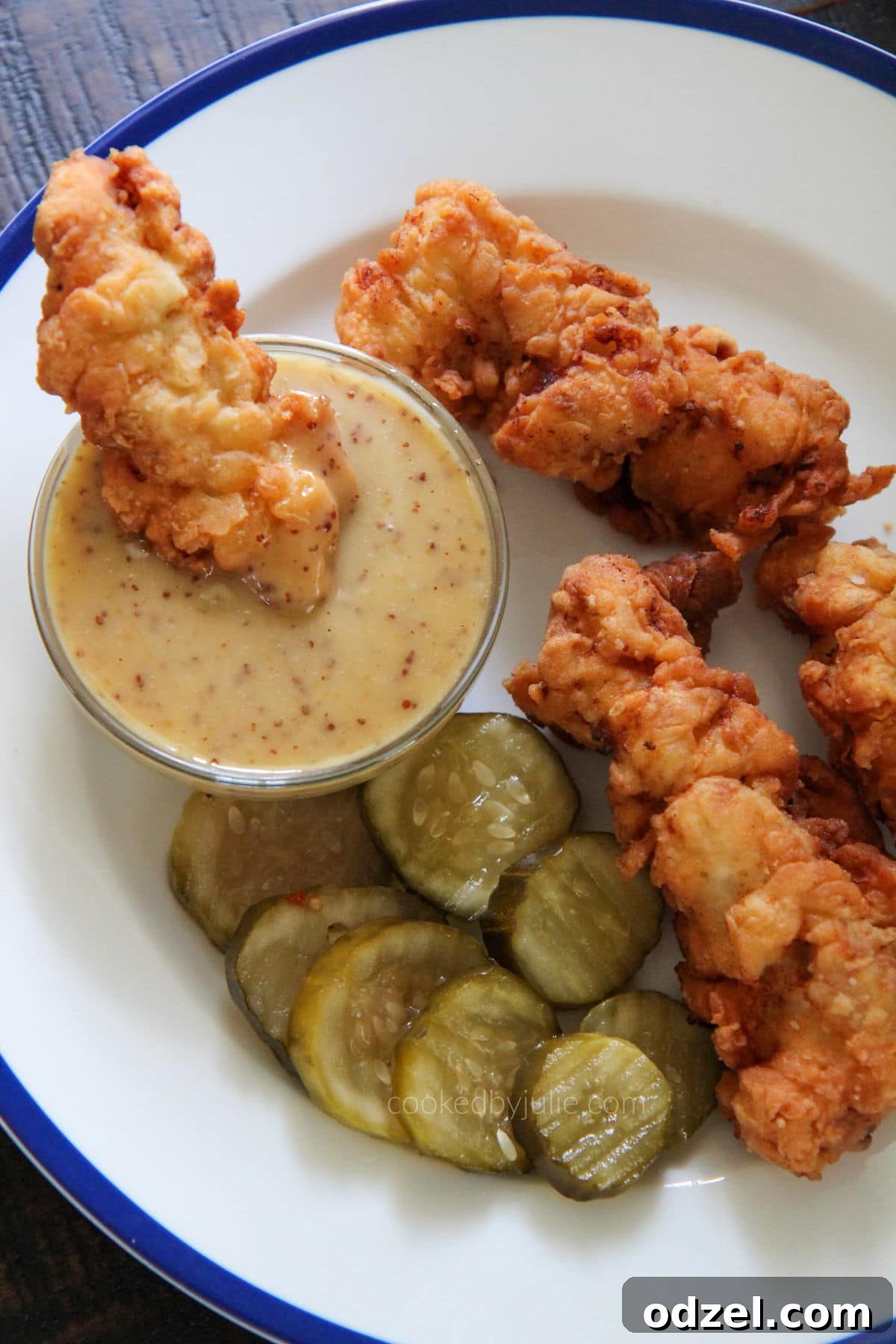 Golden-brown, crispy buttermilk chicken tenders served with pickles and a side of homemade honey mustard sauce on a white plate, ready to be enjoyed.