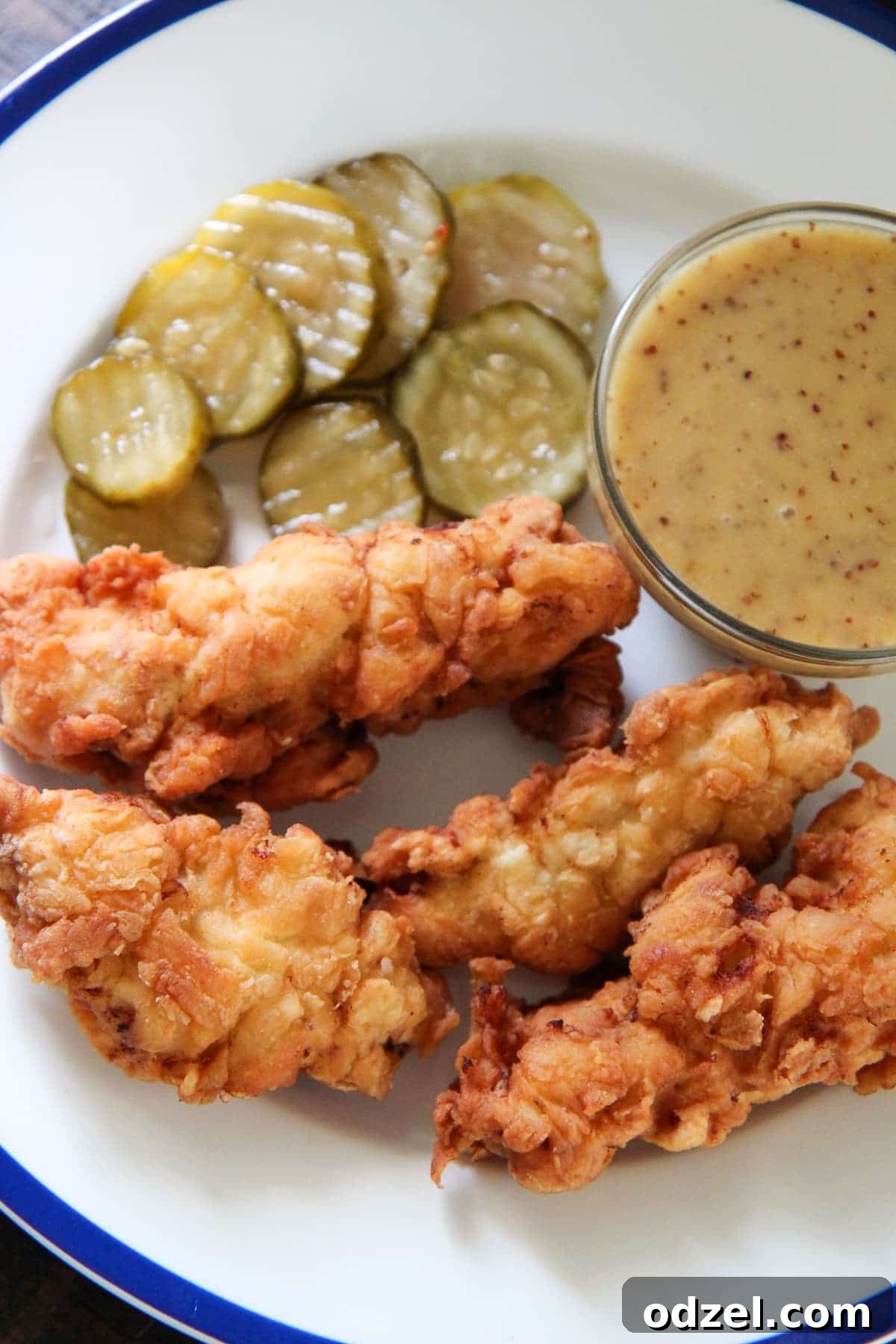 A beautifully arranged plate featuring golden-brown crispy chicken tenders, a small bowl of creamy homemade honey mustard sauce, and fresh pickle slices, emphasizing the deliciousness of the meal.