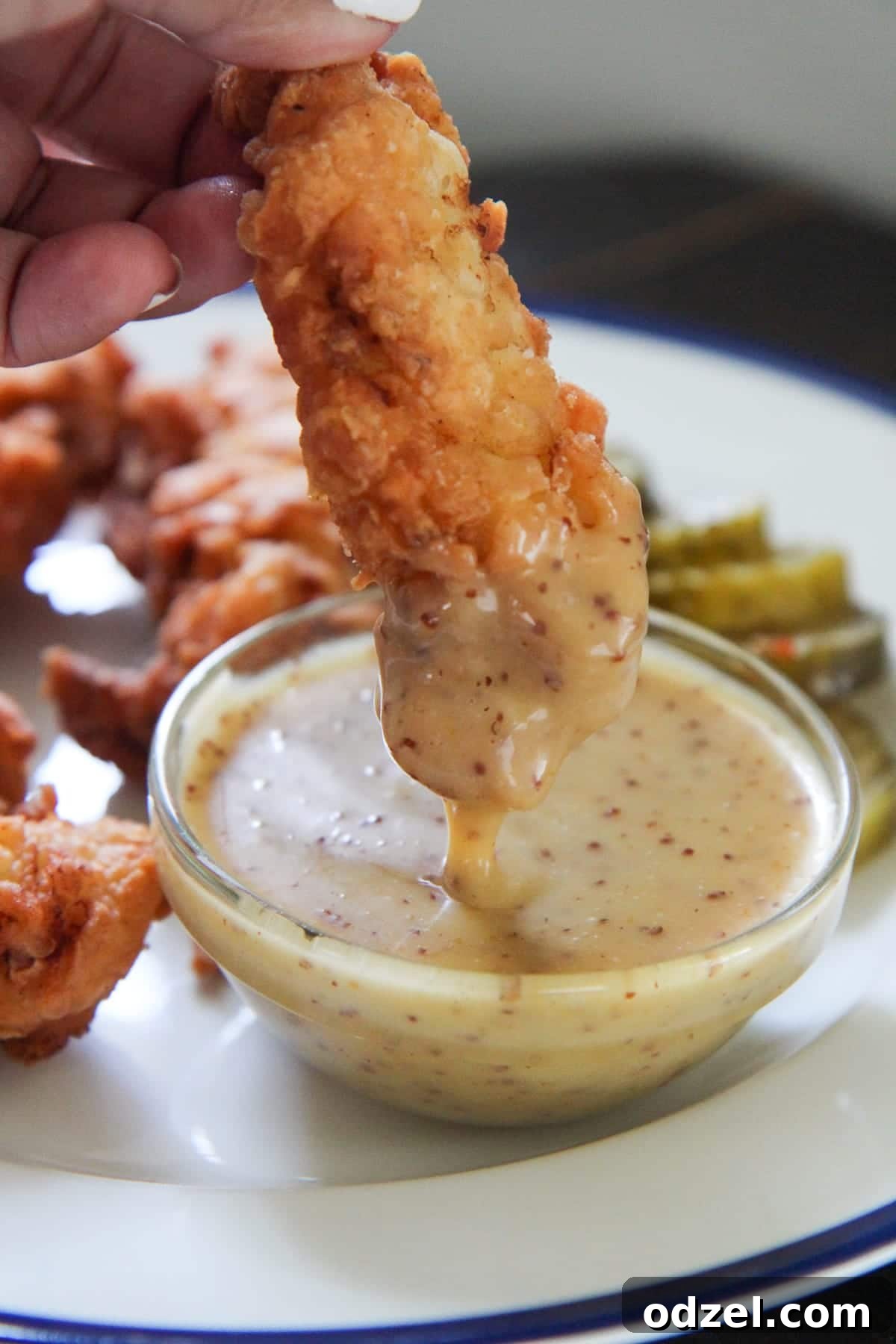 A close-up shot of a perfectly golden-brown fried chicken tender being dipped into a small bowl of creamy, homemade honey mustard sauce, highlighting the sauce's texture.
