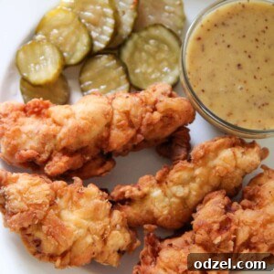 A top-down view of golden-brown crispy chicken tenders, a small bowl of creamy homemade honey mustard sauce, and fresh pickle slices on a pristine white plate.