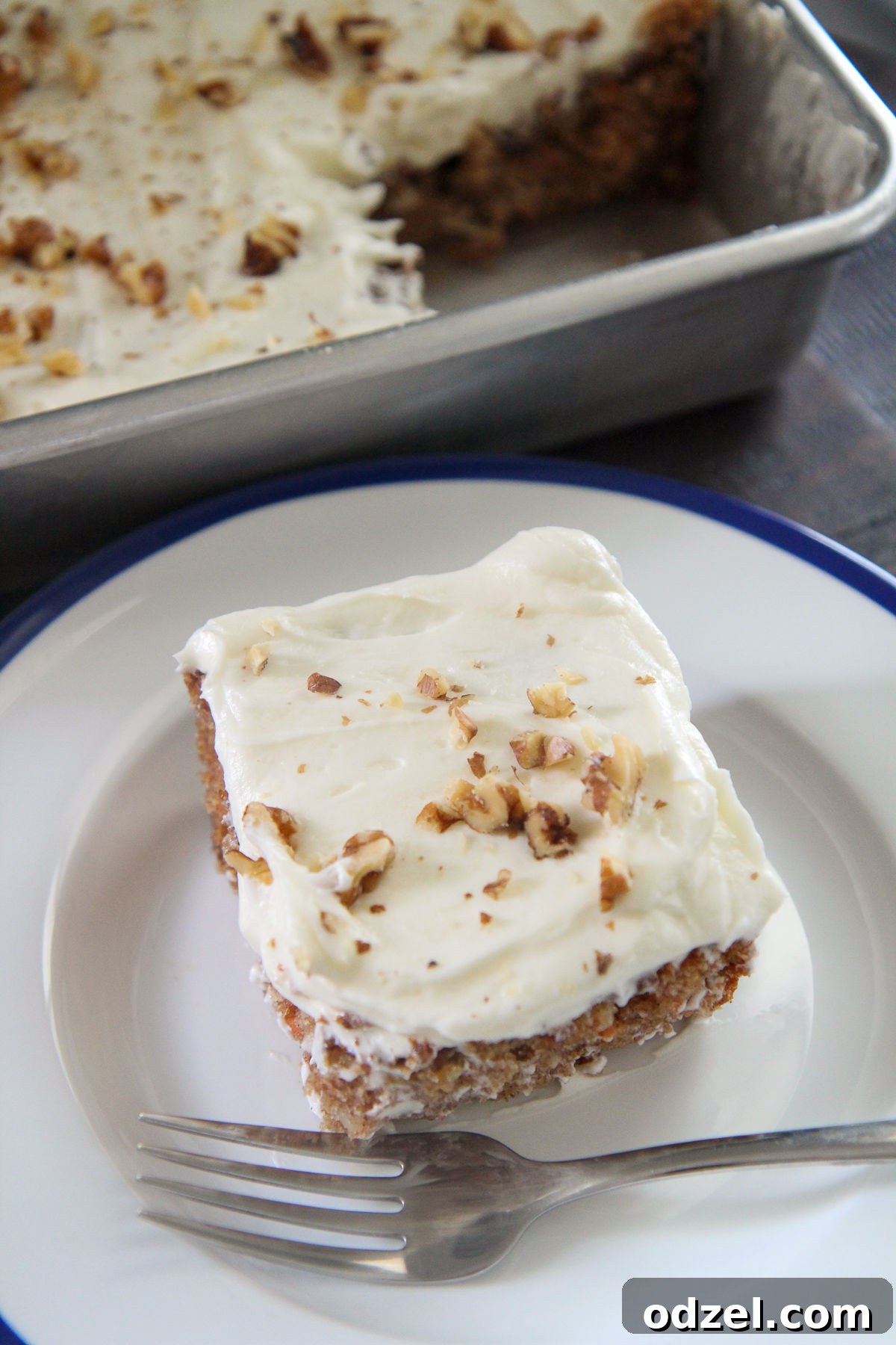 A perfectly sliced piece of moist carrot cake with thick cream cheese frosting and chopped walnuts, served on a white plate with a silver fork.