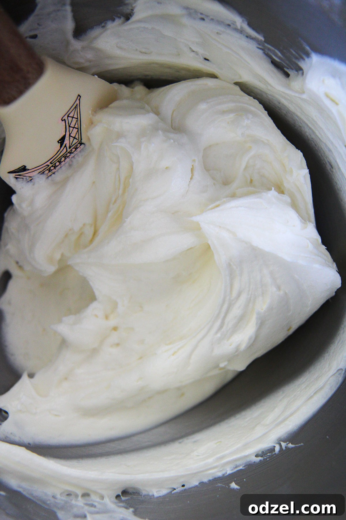 A bowl of creamy, white homemade cream cheese frosting with a rubber spatula resting inside, showing its smooth and luscious texture.