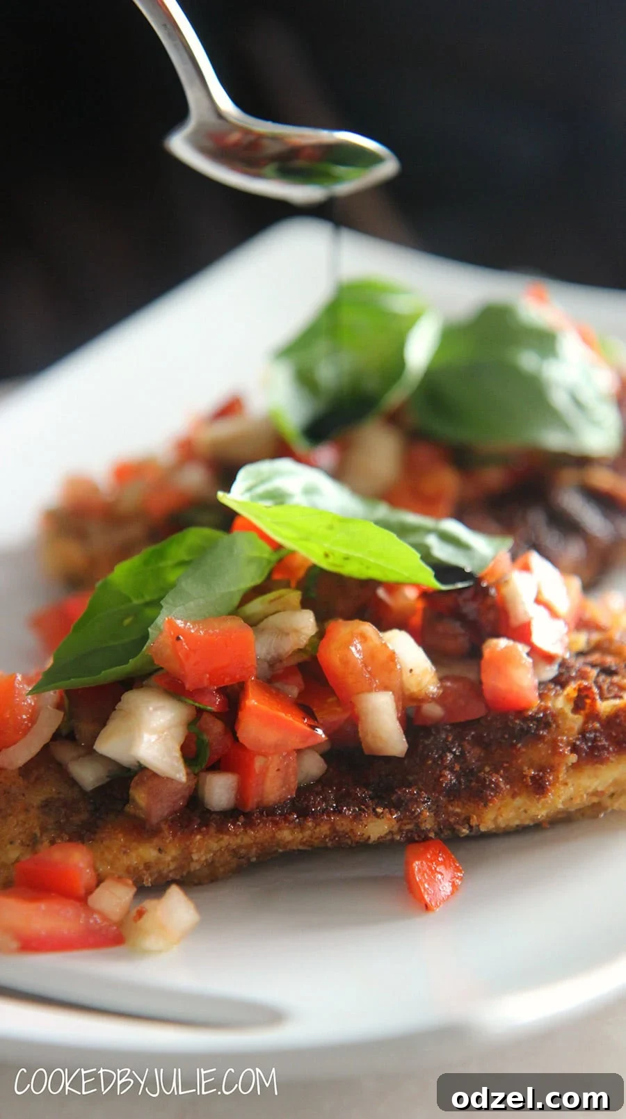 Tuscan Chicken Bruschetta 2 Golden-brown fried chicken breasts topped with a vibrant mixture of diced Roma tomatoes, red onions, and fresh basil leaves. A spoon drizzles rich balsamic glaze over the entire dish, presenting a restaurant-quality meal on a white plate.