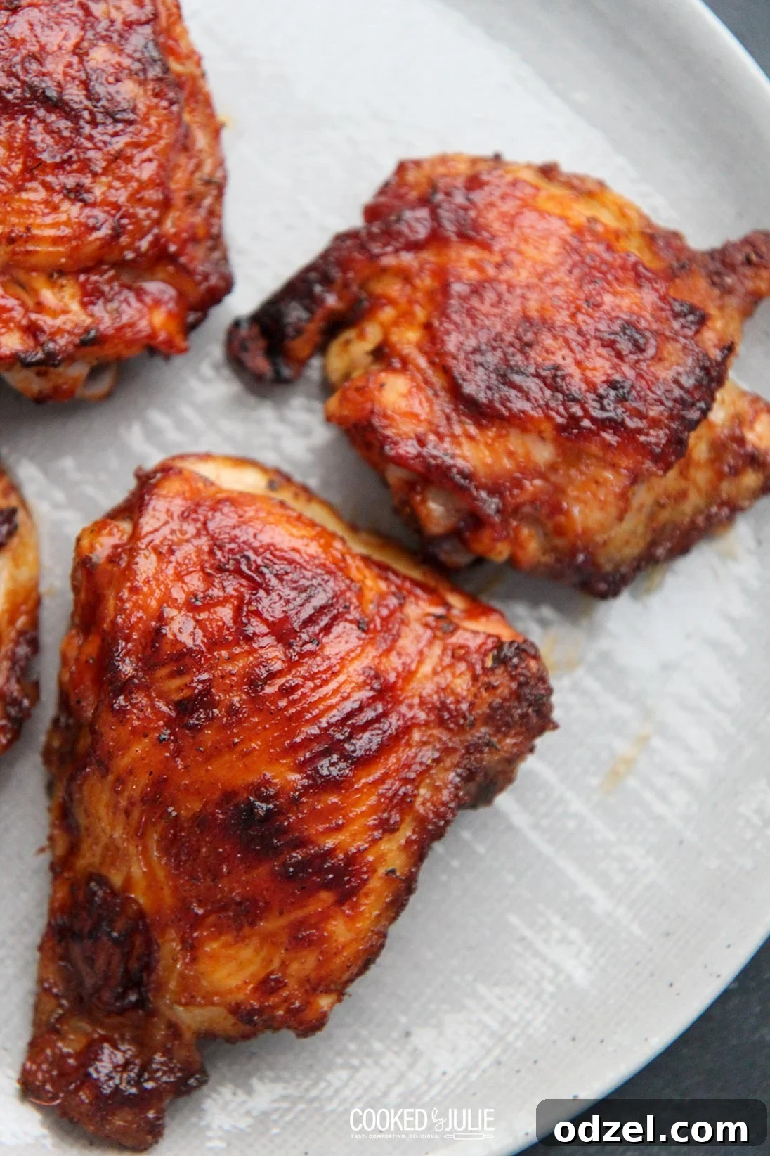 Three beautifully glazed BBQ chicken thighs on a gray serving plate, garnished with fresh herbs