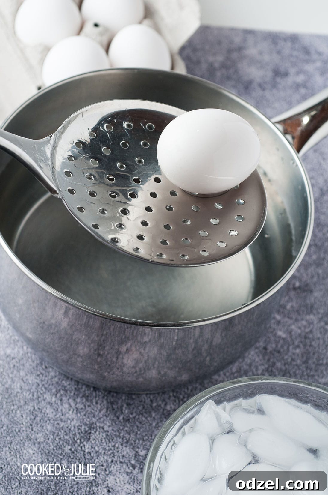 A straining spoon lifting a white egg out of a metal saucepan, preparing to transfer to an ice bath