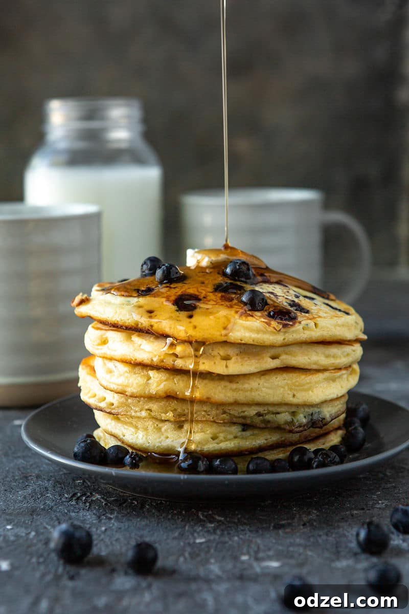 Six fluffy blueberry pancakes on a blue plate, garnished with melted butter, maple syrup, and fresh blueberries. A jug of milk and two mugs are visible in the background.
