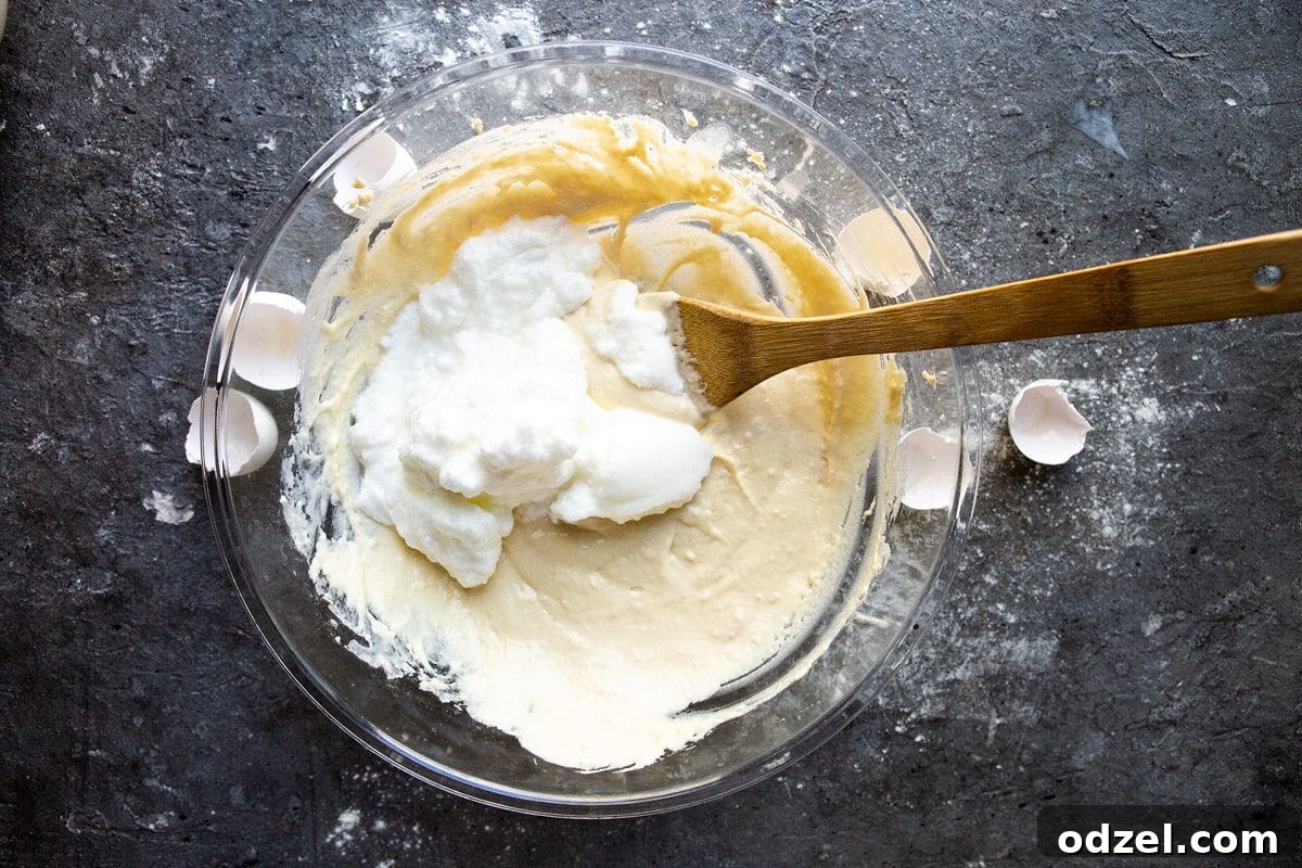 A mixing bowl containing light yellow pancake batter with freshly whipped egg whites partially folded in, alongside a rubber spatula and eggshells.
