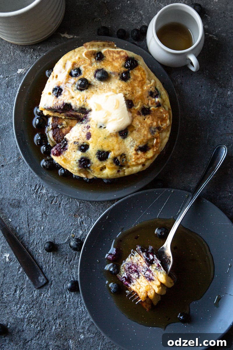 A short stack of three blueberry pancakes on a white plate, next to a smaller plate with a fork and four cut pancake slices.