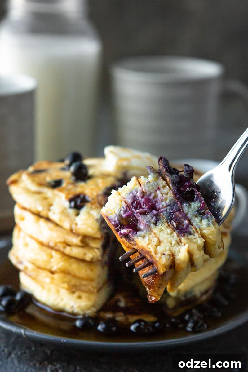 A short stack of blueberry pancakes on a plate, alongside a fork holding several pieces of pancake, ready to be eaten.