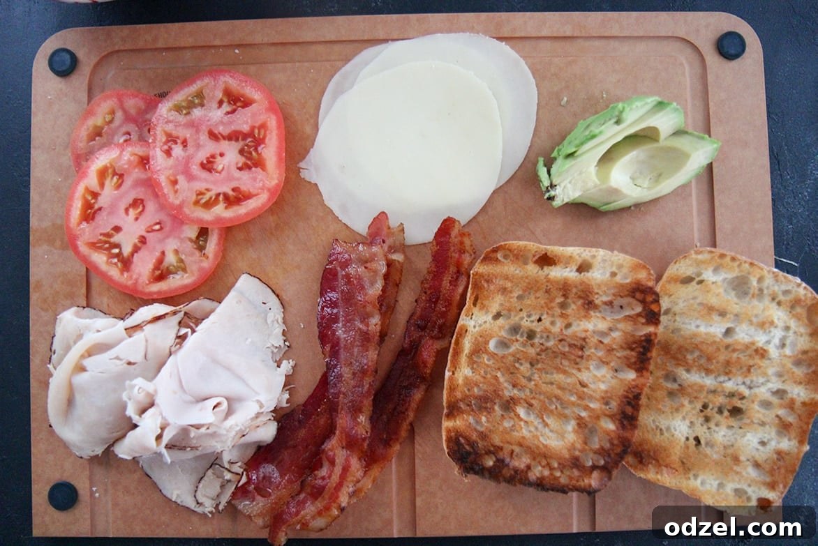 Loaded Turkey Bacon Avocado Dream 5 All fresh ingredients for a turkey bacon avocado sandwich laid out on a wooden board: sliced tomatoes, crispy bacon, deli turkey, provolone cheese, sliced avocado, and ciabatta bread.