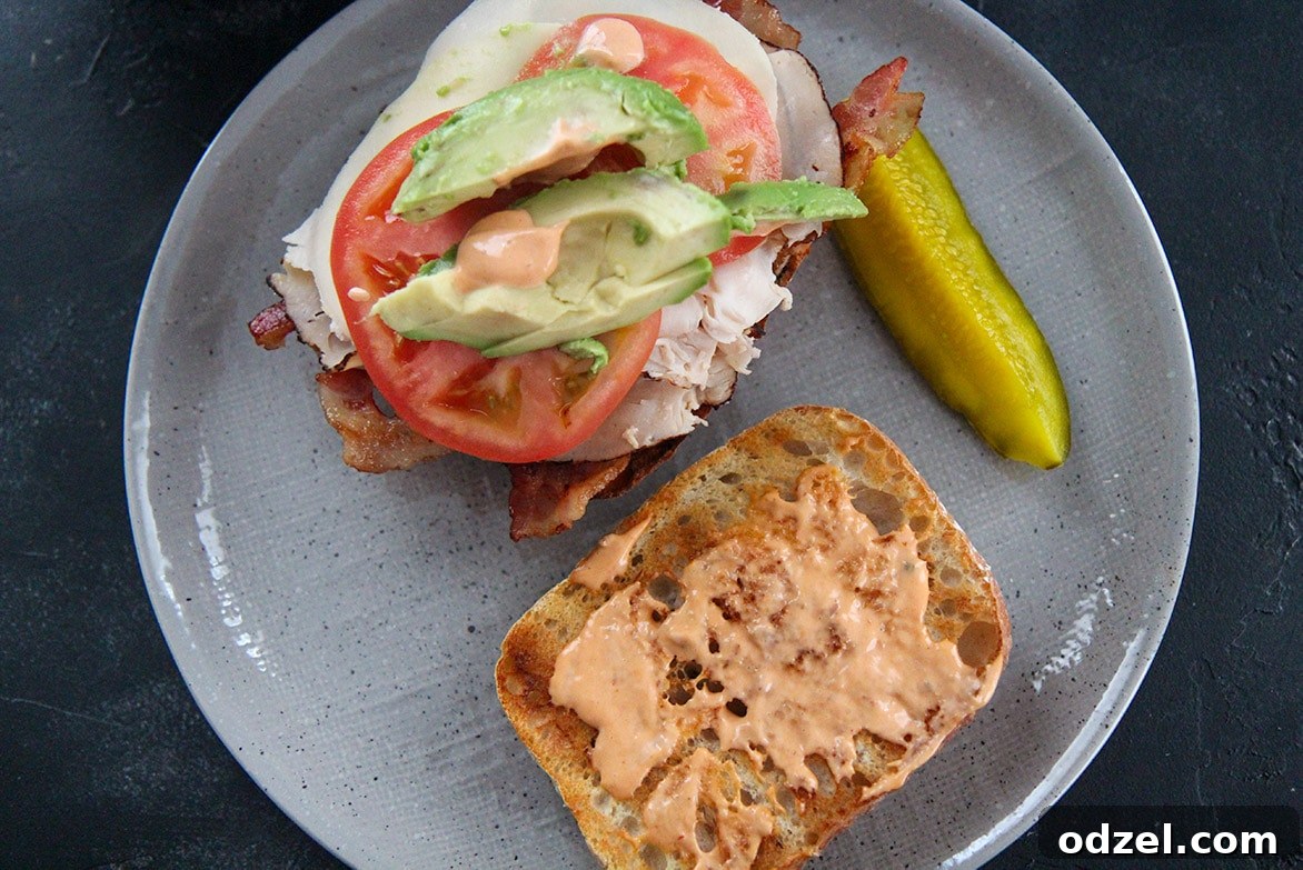 Loaded Turkey Bacon Avocado Dream 6 A perfectly assembled turkey bacon avocado sandwich with a dill pickle spear, presented on a minimalist gray plate.