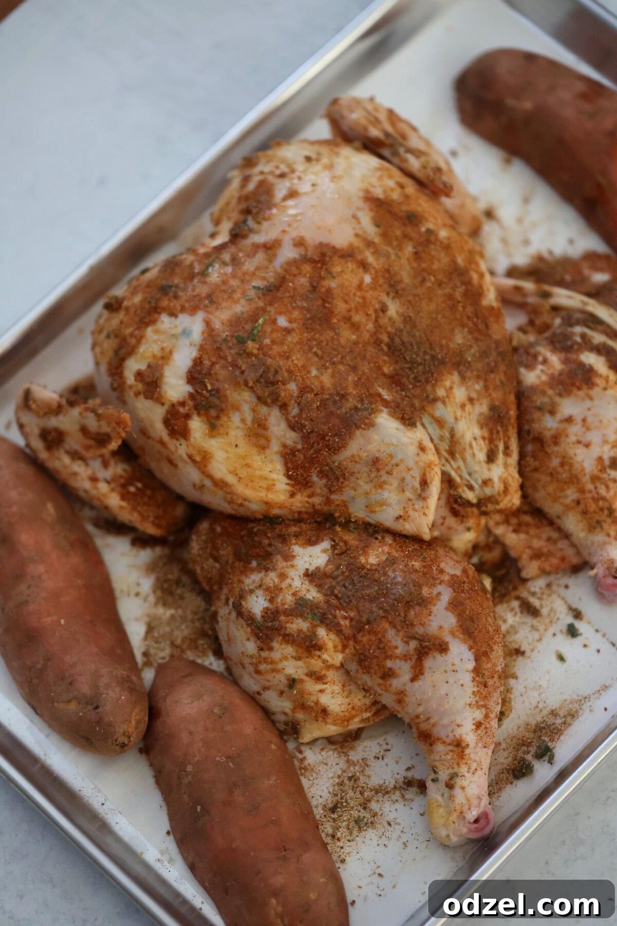 Raw seasoned spatchcock chicken and uncooked sweet potatoes arranged on a sheet pan, ready for roasting.