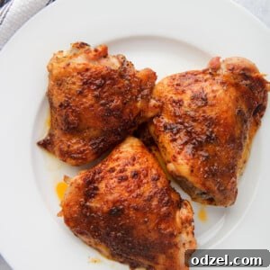 Three baked chicken thighs on a white plate with a gray and white towel on the side.