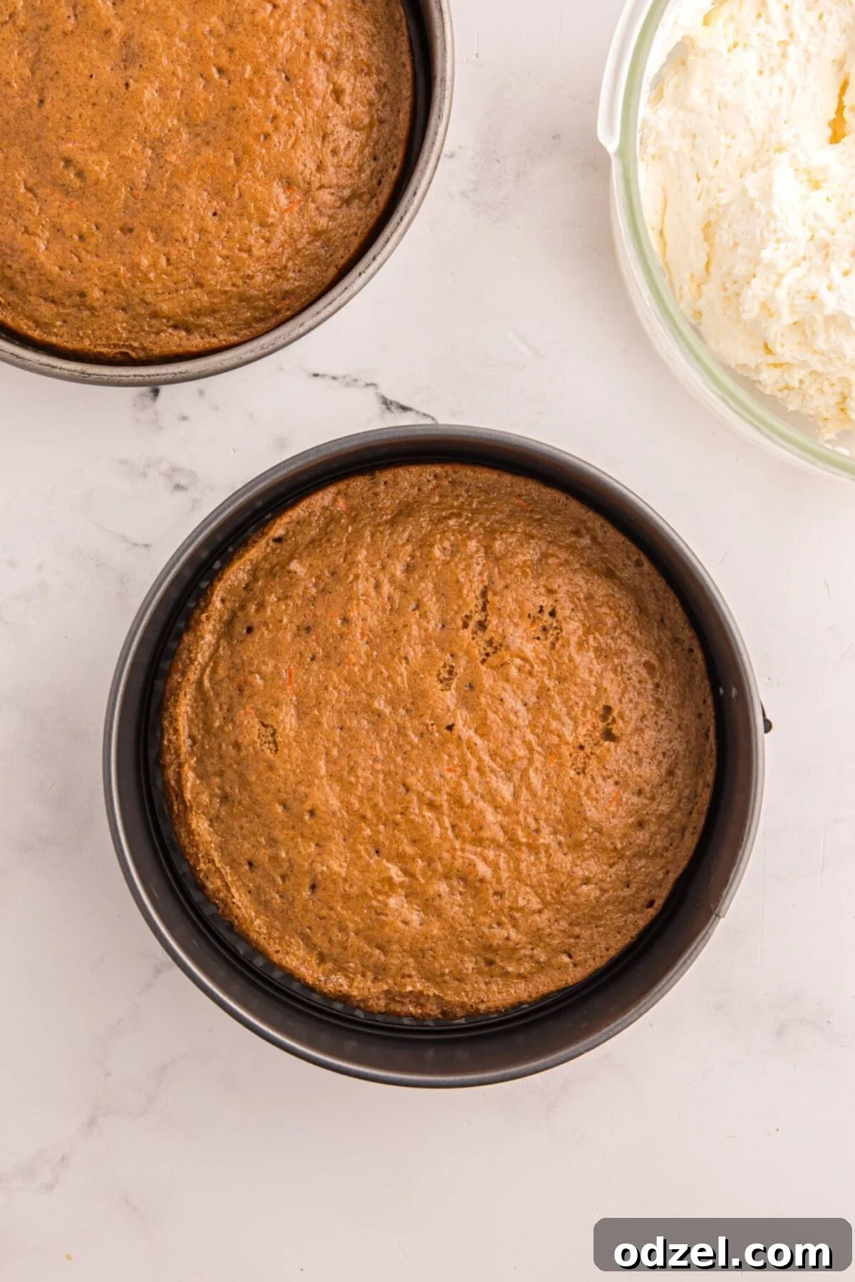 Two baked carrot cake layers in round cake pans with cream cheese frosting in a bowl.