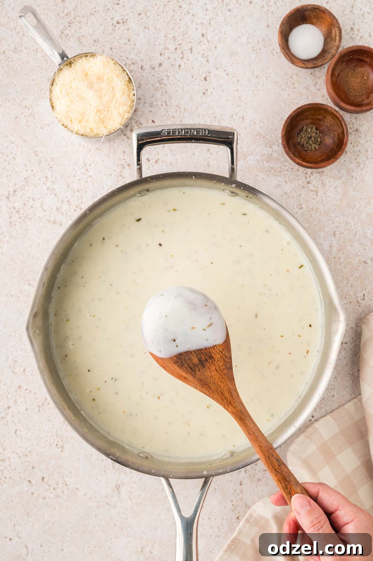 Creamy Pesto Chicken Lasagna Recipe 7 White sauce coating the back of a wooden spoon, with parmesan cheese and seasonings beside the skillet.