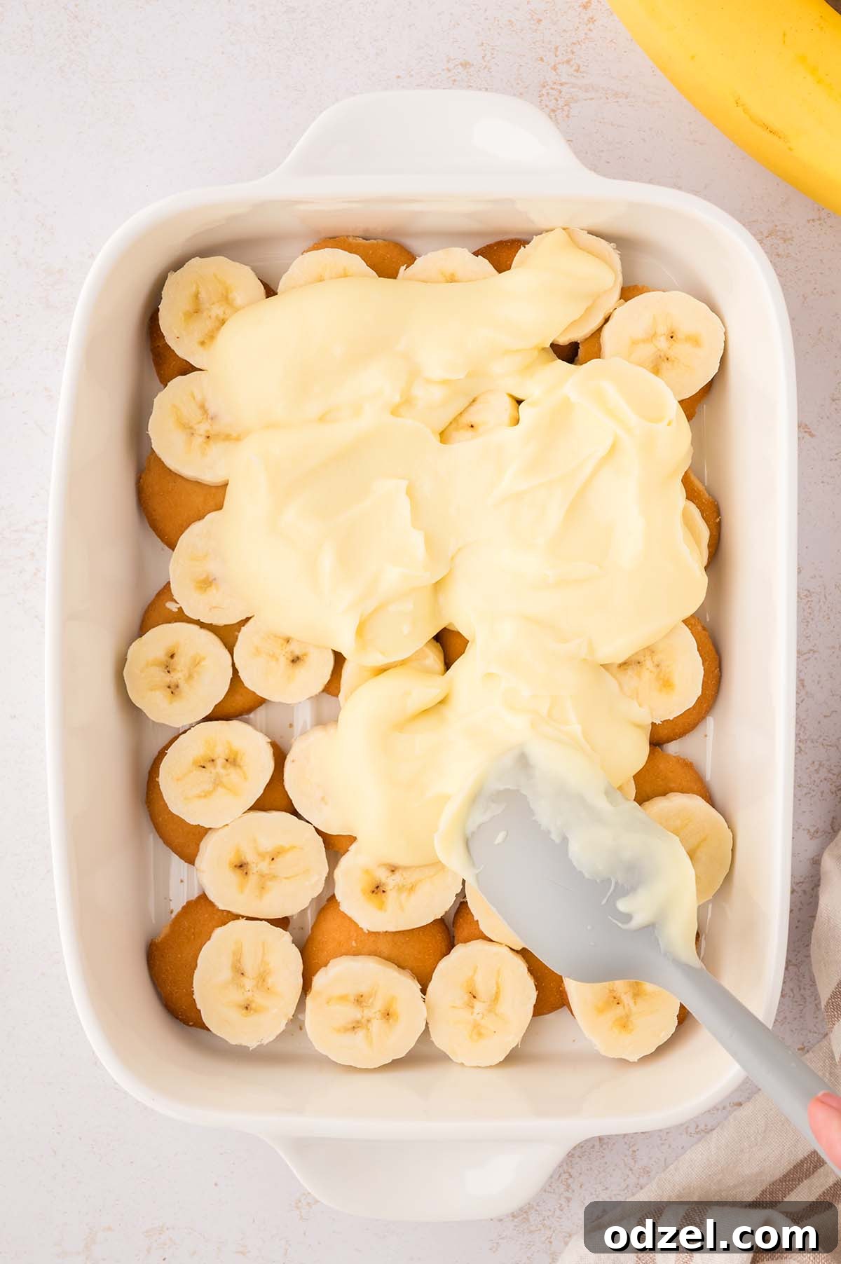 Southern Classic Banana Pudding Recipe 8 yellow pudding being spread on top of banana slices and vanilla wafers.