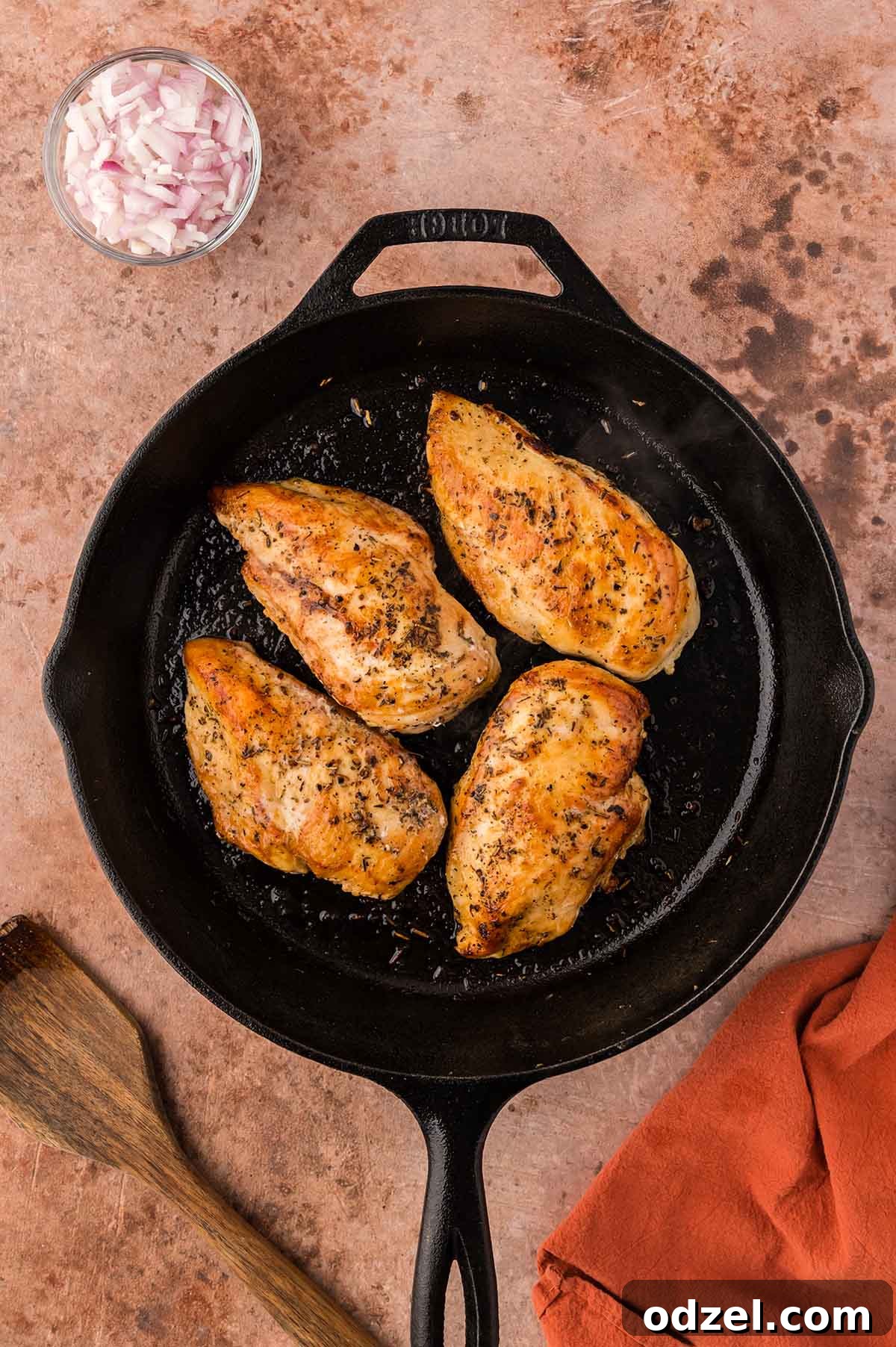 four chicken breasts cooking in an iron skillet with shallots on the side.