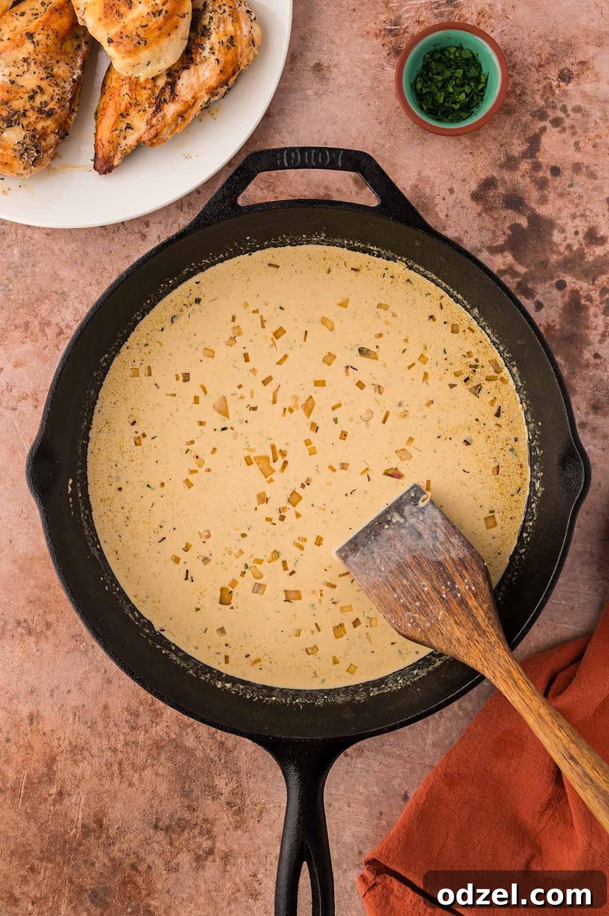cream sauce in a skillet with a wooden spoon and chicken and parsley on the side.